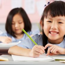 Smiling young girl in a classroom with a pencil and workbook. Other children doing schoolwork behind her.